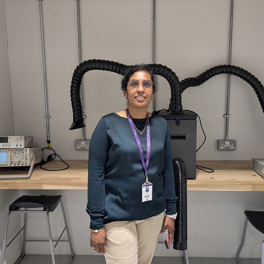 Pradeepa Sundaram, Engineering Lecturer at Richmond upon Thames College - she is stnading in front of a bench with equipment on it and large black tubes coming out of it. She is wearing glasses with a teal long sleeve top and cream trousers and a purple lanyard around her neck. 