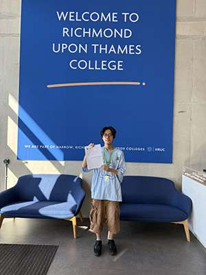 Yuto - a Richmond upon Thames College student is holding his A-Level results in his hands and standing in front of the large 'Welcome to Richmond upon Thames College' blue wall banner in reception. There are two large blue chairs behind him.
