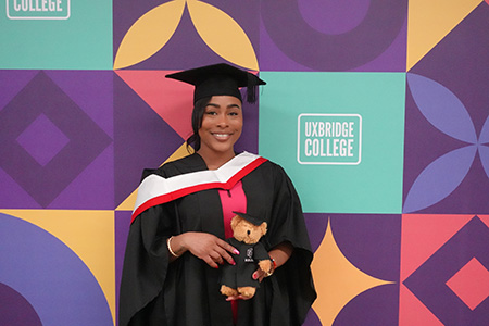 Female HRUC HE graduation student, holding a teddy bear with black graduation robes. She is wearing cap and gown with red and white on her hood. She is standing in front of the Uxbridge graphic branding on a wall.                   