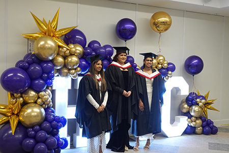 Three female HE graduates posing for a photo in front of the lit up HRUC sign with purple and gold balloons around it. They are all wearing their graduation robes - caps and gowns with red and white colours on the hood.