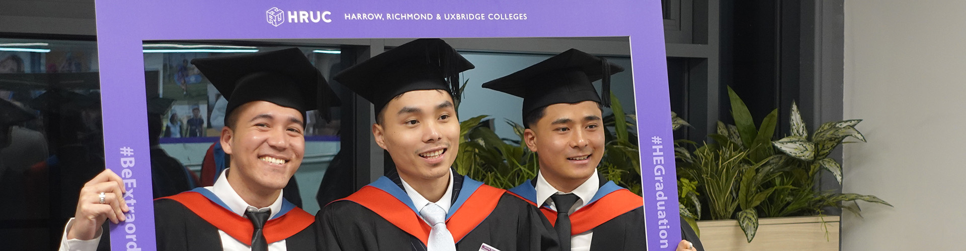 Three of our HE graduates posing for a photo holding the purple HRUC photo frame. They are all wearing their graduation robes - caps and gowns with red and blue colours. 